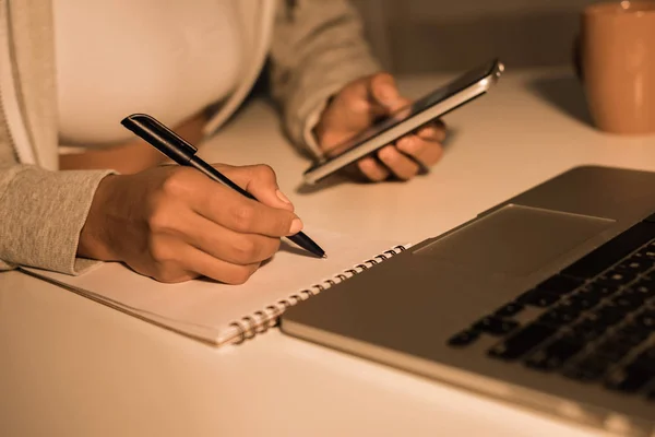 Woman writing at notebook while holding smartphone — Stock Photo