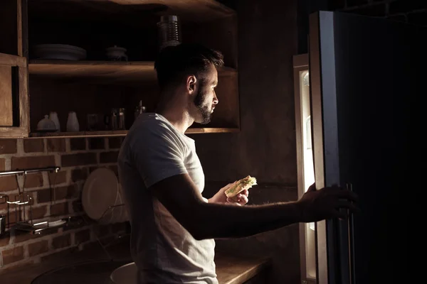 Homem de pijama comendo e olhando para geladeira — Fotografia de Stock