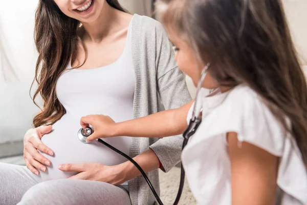 Girl listening to pregnant belly through stethoscope — Stock Photo