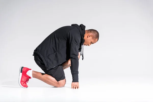 Vue latérale du jeune sportif afro-américain en sweat à capuche à genoux isolé sur gris — Photo de stock