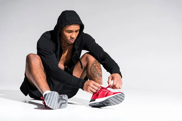 Young african american sportsman in hoodie sitting and tying shoelaces on grey — Stock Photo