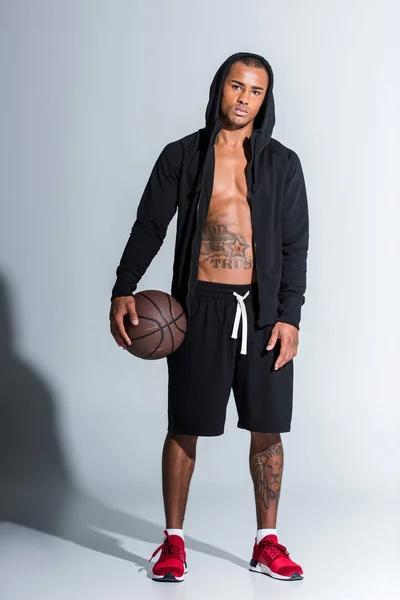 Ganzkörperansicht des sportlichen afrikanisch-amerikanischen Mannes mit Basketballkorb, der auf grau in die Kamera schaut — Stockfoto