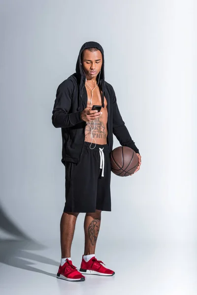 African american man with basketball ball wearing earphones and using smartphone on grey — Stock Photo