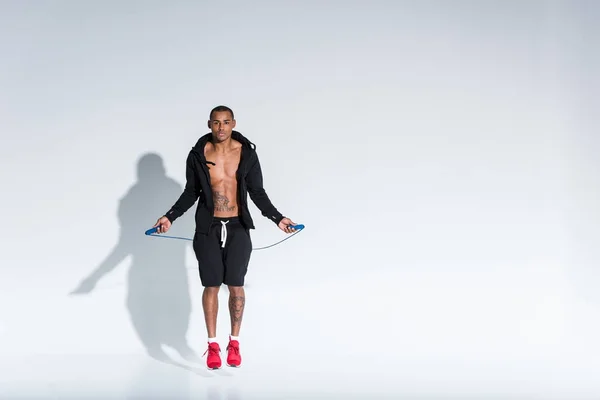 Giovane sportivo afroamericano che si allena con corda da salto e guarda la macchina fotografica sul grigio — Foto stock