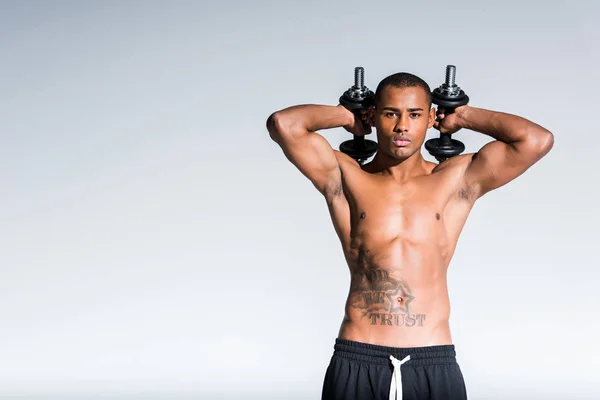 Deportista afroamericano sin camisa sosteniendo mancuernas y mirando a la cámara aislada en gris - foto de stock