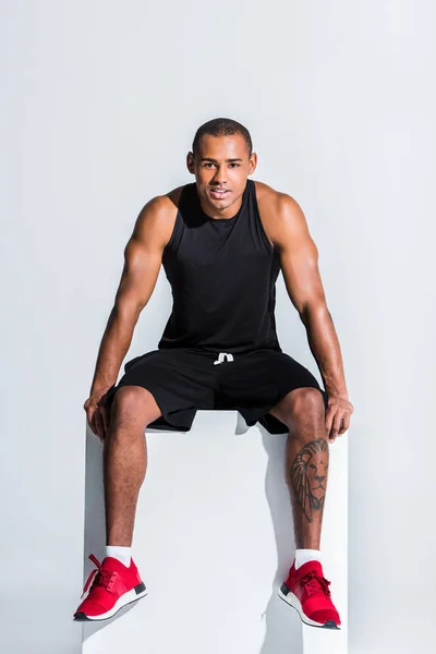 Joven afroamericano deportista en ropa deportiva sentado y sonriendo a la cámara en gris - foto de stock