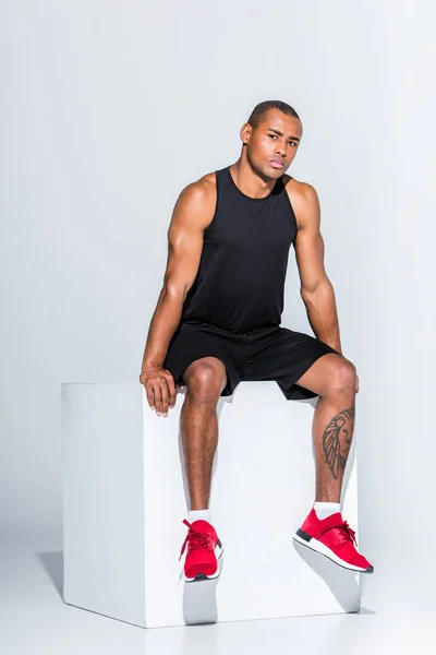 Ganzkörperansicht des jungen afrikanisch-amerikanischen Sportlers, der auf grau sitzt und in die Kamera schaut — Stockfoto