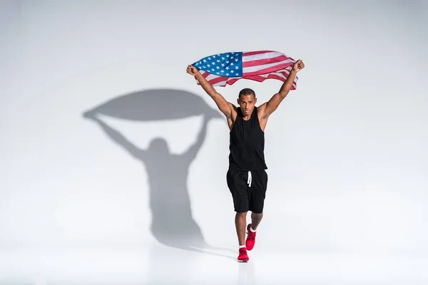 Joven afroamericano deportista con bandera americana mirando a la cámara en gris - foto de stock