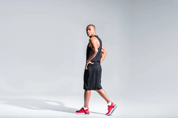Joven afroamericano deportista con las manos en la cintura caminando y mirando a la cámara en gris - foto de stock
