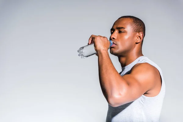 Молодой африканский американский спортсмен пьет воду из бутылки, изолированной на сером — стоковое фото