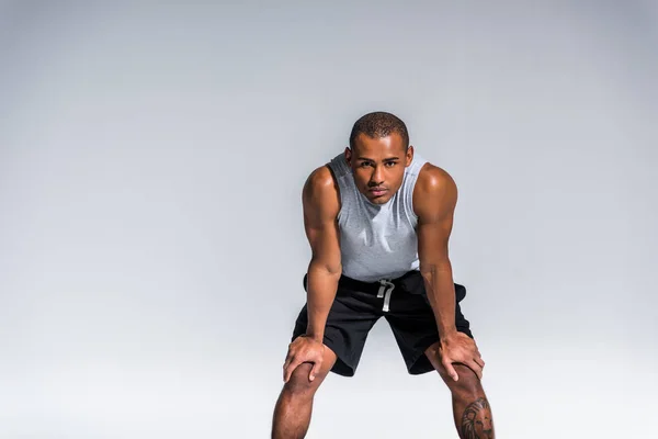 Атлетичный молодой африканский спортсмен, стоящий с руками на коленях и смотрящий в камеру на сером — стоковое фото