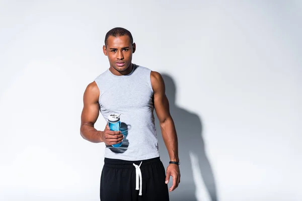 Jovem desportista afro-americano segurando garrafa de esportes com água e olhando para a câmera em cinza — Fotografia de Stock