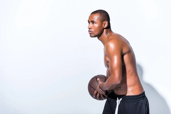 Vista lateral del hombre afroamericano sin camisa sosteniendo la pelota de baloncesto y mirando hacia otro lado en gris - foto de stock