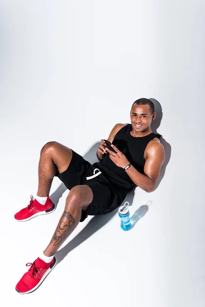 Vista de ángulo alto del apuesto deportista afroamericano usando un teléfono inteligente y sonriendo a la cámara en gris - foto de stock