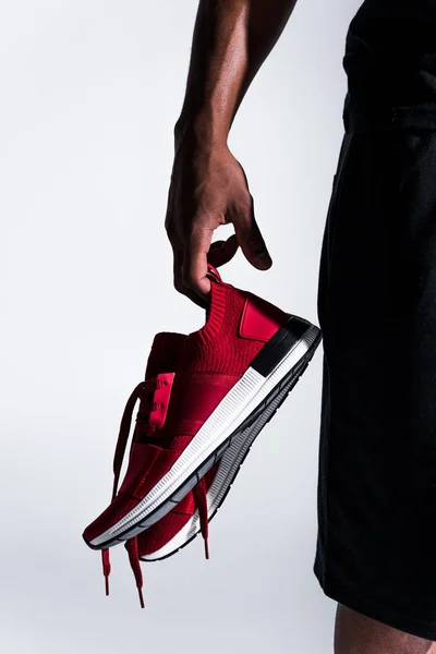 Close-up partial view of african american sportsman holding sports shoes isolated on grey — Stock Photo