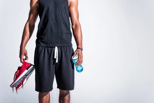 Cropped shot of young african american sportsman holding red sneakers and bottle of water isolated on grey — Stock Photo