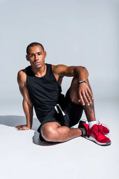 Молодой африканский американский спортсмен в фитнес-трекере и смотрит в камеру на сером — стоковое фото
