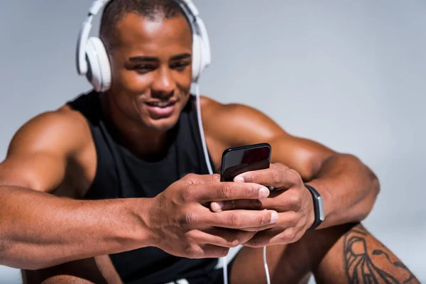 Apuesto sonriente joven afroamericano deportista en auriculares usando smartphone - foto de stock