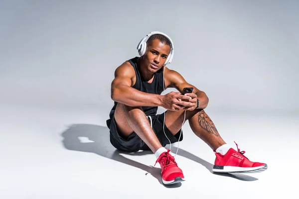 Junger gutaussehender afrikanisch-amerikanischer Sportler mit Kopfhörer sitzt und Smartphone auf grau — Stockfoto