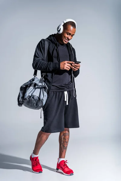 Молодий усміхнений афроамериканський спортсмен в навушниках за допомогою смартфона після тренування на сірому — стокове фото