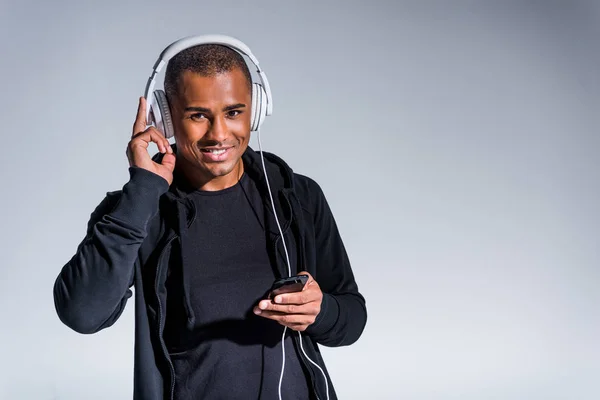 Joven afroamericano hombre en auriculares usando teléfono inteligente y sonriendo a la cámara aislado en gris - foto de stock