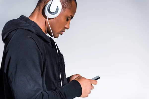 Vista lateral del joven afroamericano en audífonos usando smartphone aislado en gris — Stock Photo