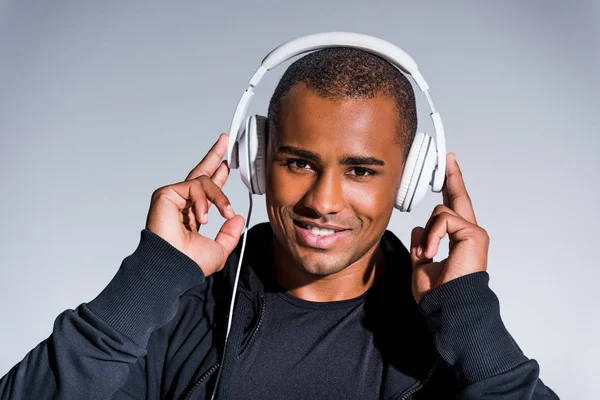 Handsome young african american sportsman listening music in headphones and smiling at camera isolated on grey — Stock Photo