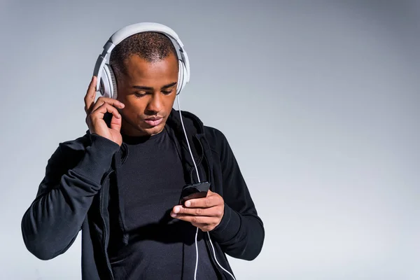Beau jeune homme afro-américain dans les écouteurs en utilisant un smartphone isolé sur gris — Photo de stock
