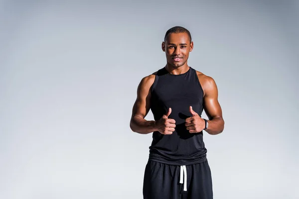 Feliz afroamericano deportista mostrando los pulgares hacia arriba y sonriendo a la cámara aislado en gris - foto de stock