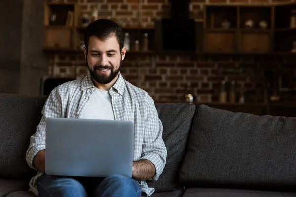 Bel homme assis sur le canapé et utilisant un ordinateur portable — Photo de stock