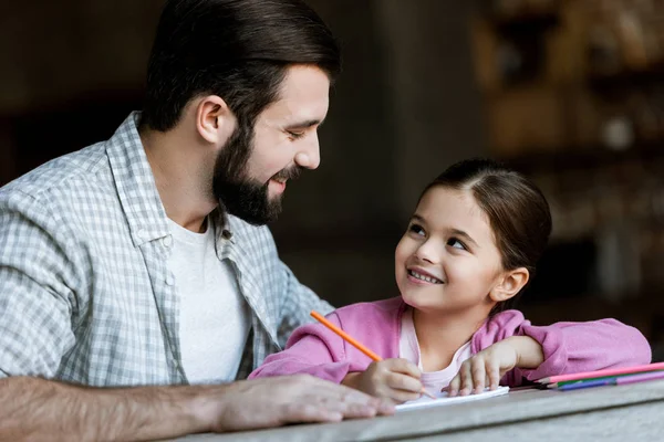 Pai feliz com a pequena filha sentada à mesa e desenho em scrapbook em casa — Fotografia de Stock