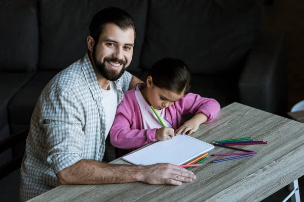 Glücklicher Vater mit kleiner Tochter am Tisch sitzend und daheim in Sammelalbum zeichnend — Stockfoto