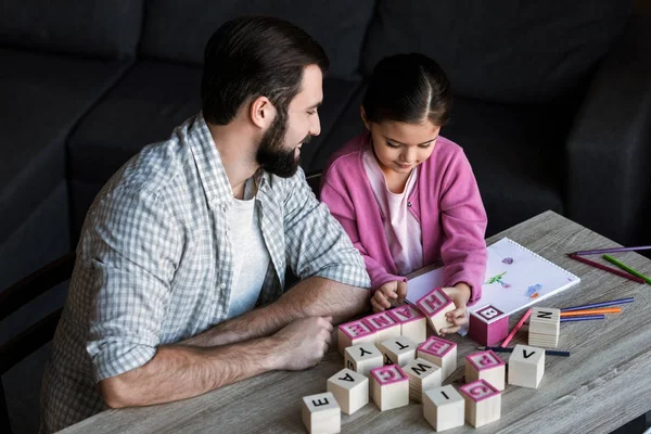 Pai com a filha sentada à mesa e fazendo palavras por cubos com letras em casa — Fotografia de Stock