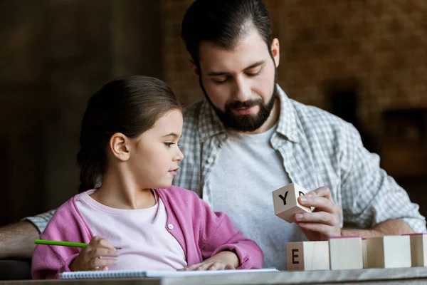 Father with daughter sitting at table and making words by cubes with letters at home — Stock Photo