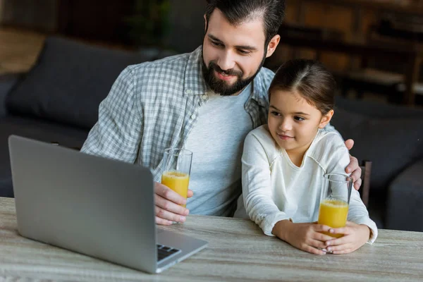 Padre sonriente con la hija sentada en la mesa con jugo y el uso de ordenador portátil - foto de stock
