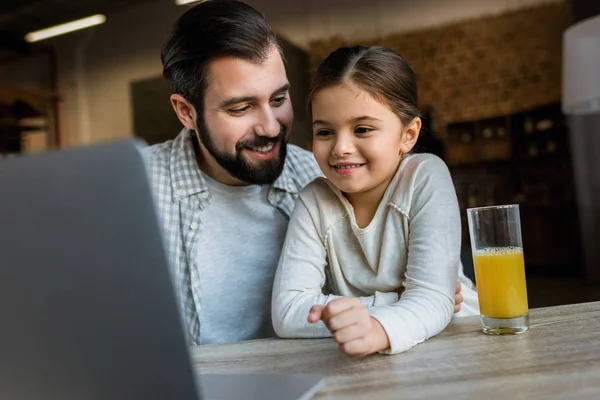 Усміхнений батько з дочкою сидить за столом з соком і використовує ноутбук — стокове фото