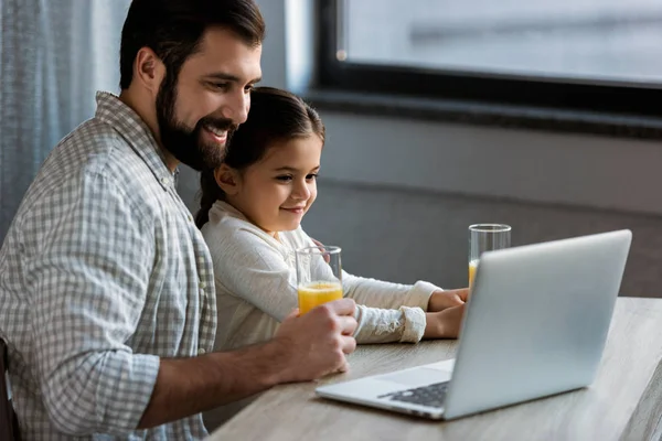 Père souriant avec sa fille assise à table avec du jus et utilisant un ordinateur portable — Photo de stock