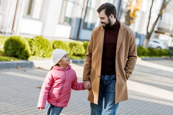 Padre alegre con hija pequeña caminando fuera en traje de otoño y mirándose. afuera - foto de stock