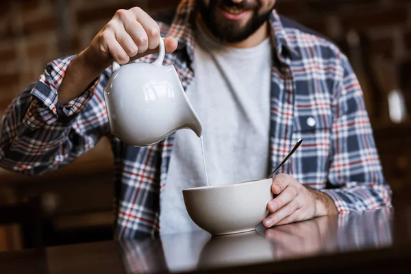 Imagen recortada de hombre vertiendo leche en un tazón con bocadillos en la cocina - foto de stock