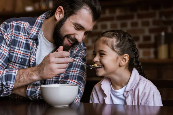 Cheerful family at table, father feeding daughter by snacks with milk at kitchen — Stock Photo