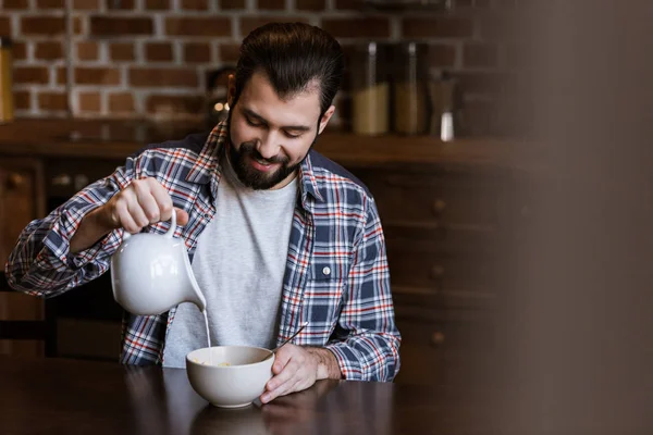 Homem bonito derramando leite em tigela com lanches na cozinha — Fotografia de Stock