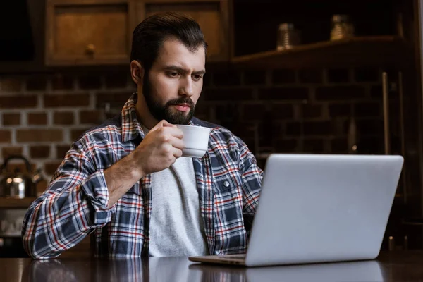 Bel homme avec tasse de café assis à table avec ordinateur portable — Photo de stock