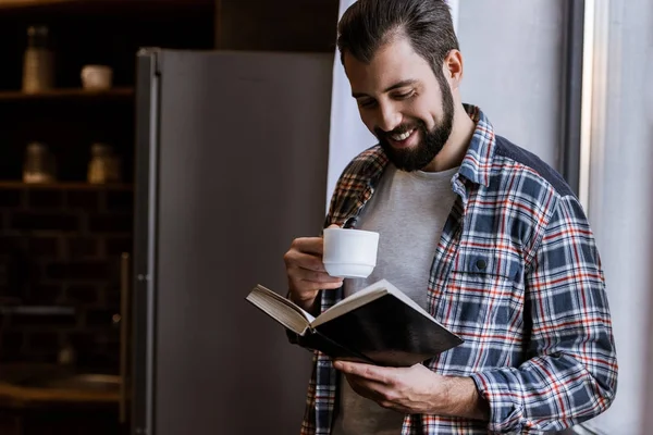 Hombre alegre con taza de café de pie junto a la ventana y libro de lectura - foto de stock