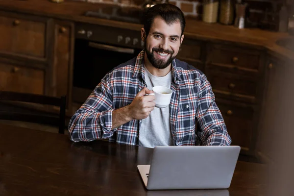 Schöner Mann mit Kaffeetasse sitzt am Tisch mit Laptop — Stockfoto