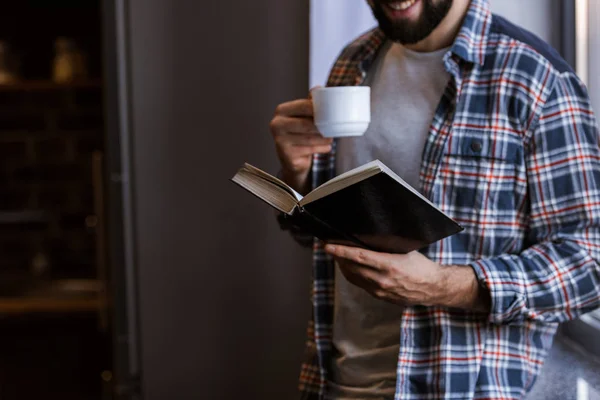 Immagine ritagliata di uomo allegro con tazza di caffè in piedi accanto alla finestra e libro di lettura — Foto stock