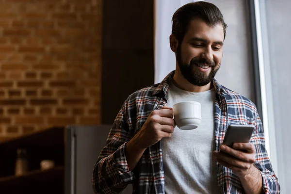 Hombre feliz sosteniendo la taza de café y el uso de teléfono inteligente en casa - foto de stock