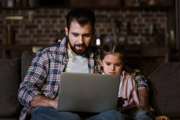 Отец с дочерью сидят на диване и пользуются ноутбуком — стоковое фото