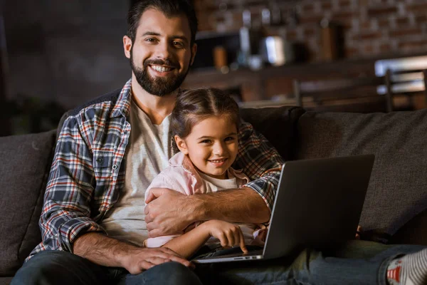 Padre feliz con hija abrazándose en el sofá y el uso de la computadora portátil - foto de stock