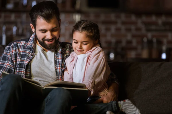 Pai feliz com a filha abraçando no sofá e lendo livro em casa — Fotografia de Stock