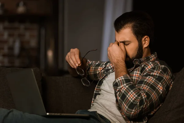 Tired man with glasses sitting on couch with laptop and rubbing head — Stock Photo